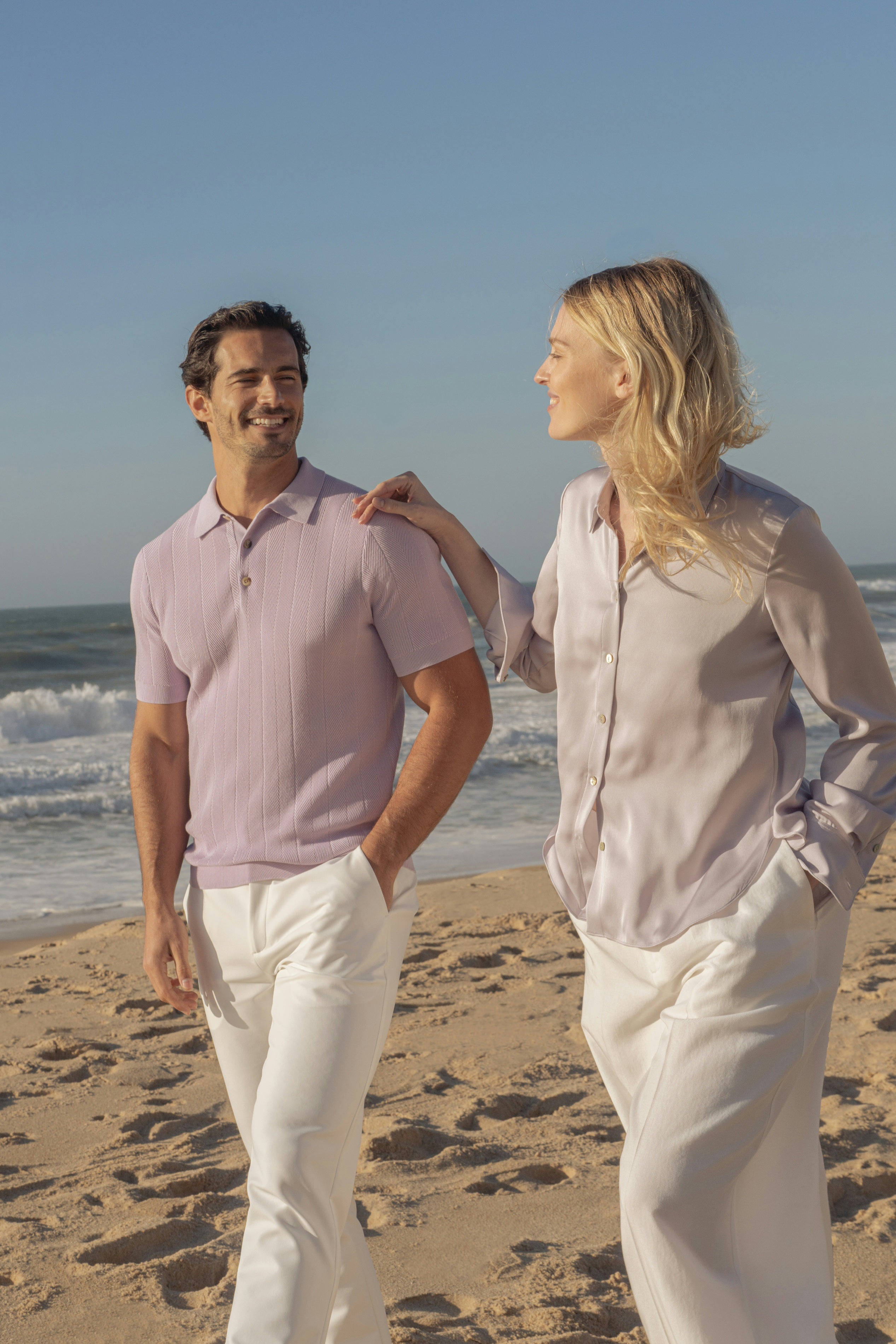 Man and woman standing on a beach, both wearing light-colored clothing.