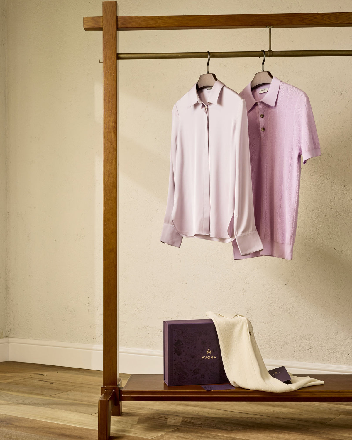 Two shirts hanging on a wooden rack with a box and towel below against a beige wall.